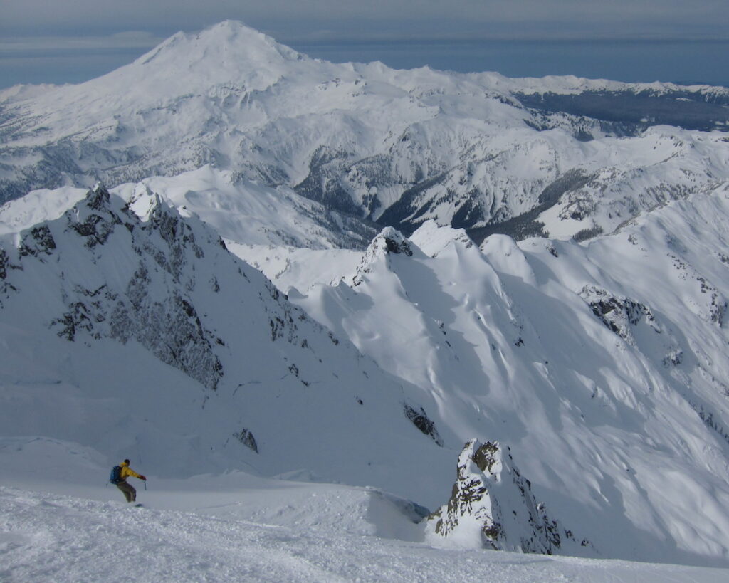 Snowboarding into the entrance of the Northwest Couloir of Mount Shuksan with  Mount Baker in the distance